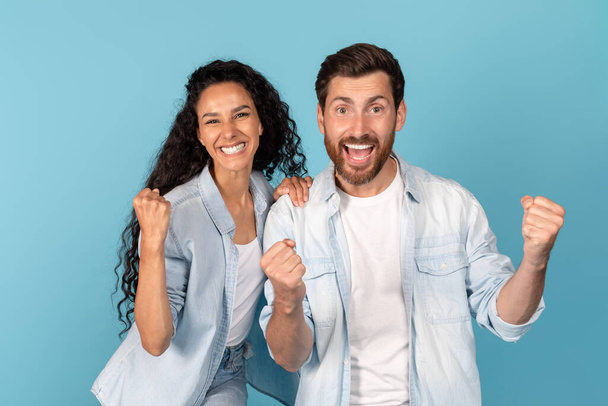 Portrait of satisfied young arab lady and european male with beard looking at camera, show victory and success sign with hands, isolated on blue background, studio. Great ad and offer, win celebration - Fotó, kép
