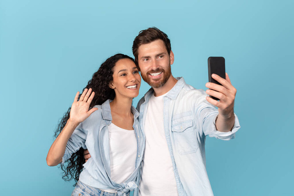 Cheerful young caucasian man with beard hugs arabic lady, waving hand, have video call on smartphone, isolated on blue background, studio. Meeting remotely, great ad and offer, new normal, covid-19 - Fotografie, Obrázek