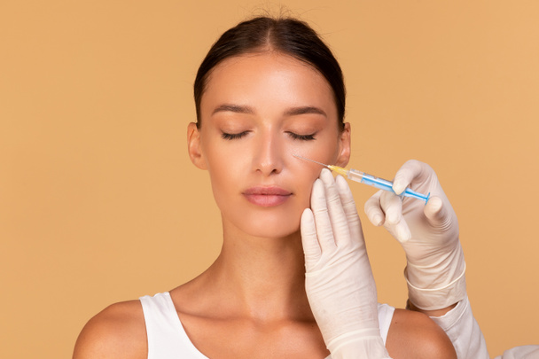 Plastic surgery and beauty injection concept. Attractive young woman receiving botox for wrinkles, standing over beige studio background. Beautician doctor holding syringe - Photo, Image