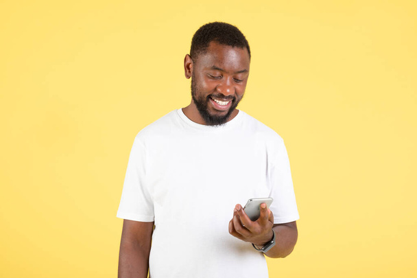 Mosolygó fekete ember telefonolvasó üzenetet használ a közösségi média alkalmazás, a kommunikáció és a szöveges Online álló sárga háttér a stúdióban. Gadgets és kommunikáció - Fotó, kép