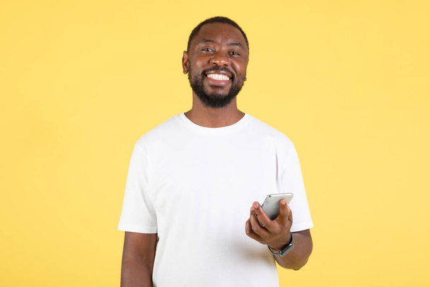 Pozytywny czarny facet za pomocą Smartphone SMS na stojąco pozowanie na żółtym tle Studio. Facet smsuje przez telefon i uśmiecha się do kamery. Świetna oferta mobilna Reklama - Zdjęcie, obraz
