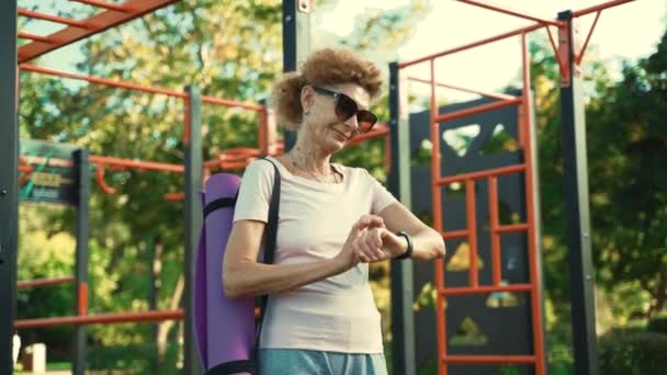 Donna anziana con tappetino yoga si trova sulla palestra di strada e utilizza orologio sportivo. Una donna anziana che tiene un blocco yoga e controlla il suo orologio da polso dopo l'allenamento. gadget sportivi moderni, concetto di attrezzature per il fitness. - Filmati, video