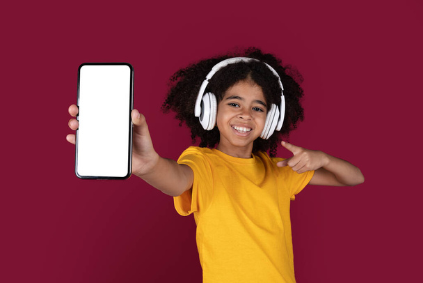 Fajna ładna czarna dziewczyna z ładnymi krzaczastymi włosami nastolatka wskazując na nowy smartfon z białym, czystym ekranem i uśmiechając się, za pomocą bezprzewodowych słuchawek, słuchając muzyki na burgundowym tle, makieta - Zdjęcie, obraz