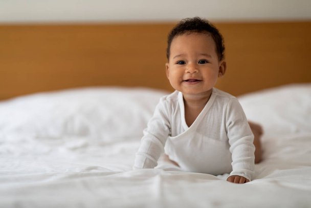 Porträt eines entzückenden schwarzen Neugeborenen, das es sich zu Hause im Bett gemütlich macht, süßes Lächeln eines afrikanischen Säuglings, das auf einem Bettlaken krabbelt, kleiner Junge oder Mädchen, das im gemütlichen Schlafzimmer liegt, Kopierraum - Foto, Bild