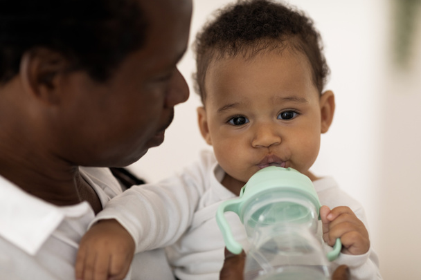 Urocze małe czarne dziecko pijące wodę z butelki relaksując się w rękach ojców, zbliżenie Strzał spragnionego słodkiego Afroamerykańskiego dziecka sączącego zdrowego drinka i patrzącego przed kamerą - Zdjęcie, obraz