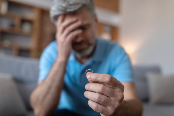Despaired sad middle aged european man suffering from divorce, holds ring in hand, blurred, copy space. Loneliness, relationship problems, breakup, stress and emotions at home during covid-19 pandemic - Foto, immagini