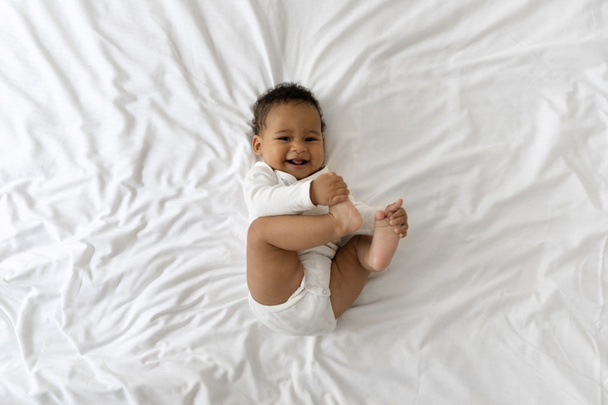 Симпатичный веселый маленький чернокожий малыш, веселящийся, лежа на кровати дома, веселый афроамериканский мальчик, играющий ногами и смеющийся во время отдыха в отеле, вид сверху, пространство для копирования - Фото, изображение