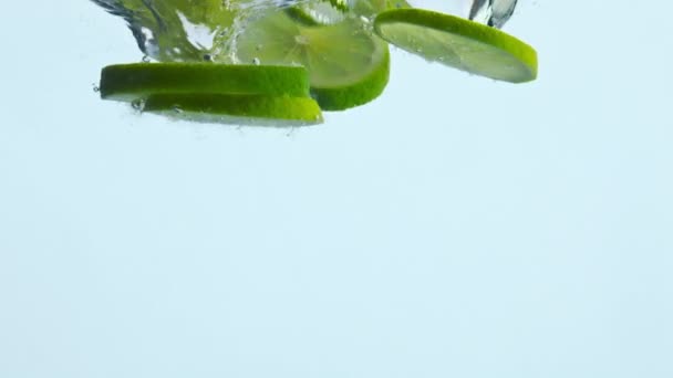 Τροπικό φέτες ασβέστη έπεσε σε κρυστάλλινο νερό λευκό φόντο από κοντά. Κομμάτια ζουμερά εσπεριδοειδή πιτσιλίζουν διαυγές υγρό σε εξαιρετικά αργή κίνηση. Φρέσκο βιολογικό συστατικό για χυμό κοκτέιλ λεμονάδα. - Πλάνα, βίντεο