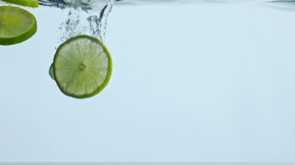 Closeup zure heldere limoenschijfjes onder water met belletjes op witte achtergrond. Cijfers sappige citrusvruchten vallen diep onder helder water in super slow motion. Vitamine smakelijk ingrediënt voor natuurlijke drank. - Video