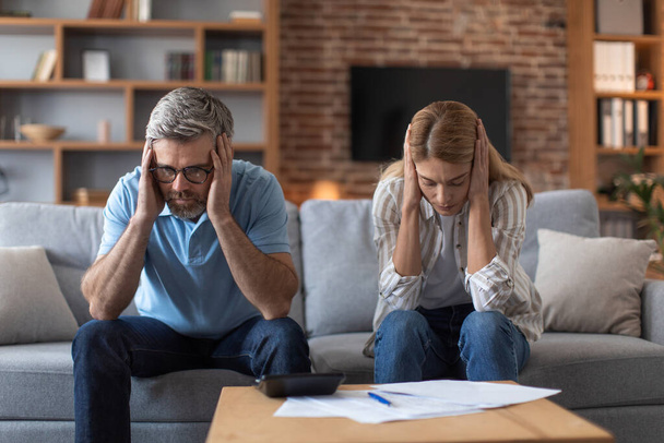 Despaired sad middle aged european couple suffer from bills in living room interior. Global crisis, stress and paperwork, bankruptcy, paying mortgage and taxes, bookkeeping, relationship problems - Foto, Imagem