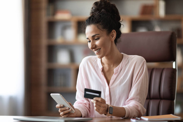Счастливая молодая женщина, сидящая в Интернете во время перерыва в работе, пользующаяся смартфоном и безграничной пластиковой кредитной картой, покупающая вещи онлайн во время работы, копирует пространство. Концепция электронной коммерции - Фото, изображение
