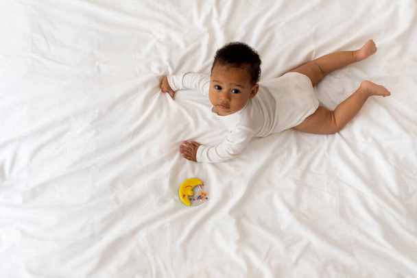 Liebenswertes kleines schwarzes Baby neben seinem Rasselspielzeug auf dem Bett liegend, neugieriges süßes afroamerikanisches Säugling auf weißer Bettwäsche im Schlafzimmer liegend und in die Kamera blickend, Draufsicht mit Kopierraum - Foto, Bild