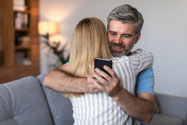 Cheerful middle aged european man hugging woman and typing on smartphone in living room interior, copy space. Relationships problems, cheating, gadget and game addiction at home due covid-19 pandemic - Photo, image