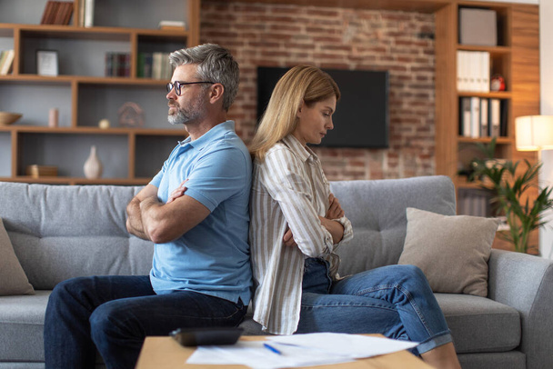 Offended sad middle aged european lady ignores man after quarrel over bills in living room interior. Crisis, work with documents, bankruptcy, payment of taxes, mortgages and relationship problems - Photo, Image