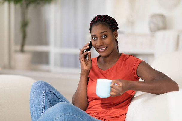 Ocio doméstico. Mujer negra joven y feliz hablando por teléfono y tomando café mientras se relaja en el sofá en casa, sonriendo mujer afroamericana descansando en un cómodo sofá en la sala de estar - Foto, Imagen