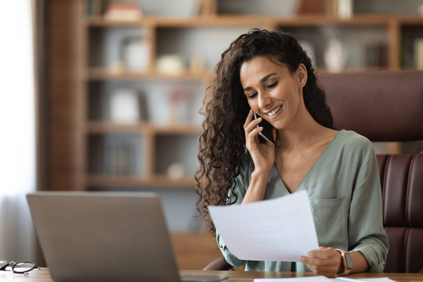 Smiling attractive millennnial businesswoman talking on phone while working on laptop, having conversation with her assistant or client, checking details, home office interior, copy space - Photo, Image