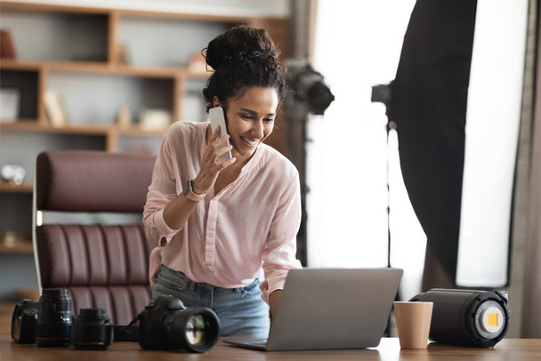 Красива творча молода жінка-фотограф, що працює в офісі, має телефонну розмову з клієнтом, дивиться на екран ноутбука, встановлює зйомку, копіює простір. Креативна художниця жіночого бізнесу
 - Фото, зображення