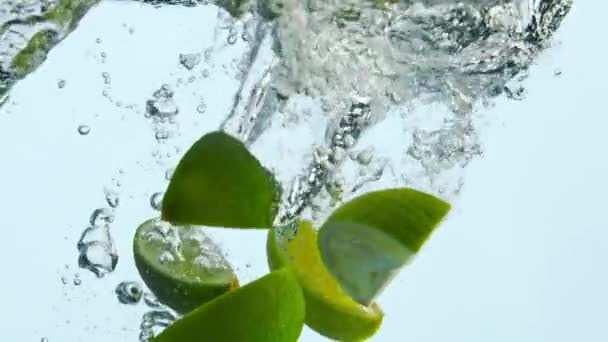Kalk wiggen vallen in helder water in super slow motion close-up. Zuurgroene citrusvruchten spatten in transparante vloeistof drijvend met belletjes. Vers smakelijk fruit ondergedompeld in kristalaquarium. Gezond voedsel. - Video