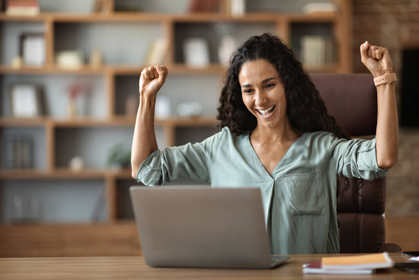 Emotional beautiful young woman entrepreneur celebrating successful business deal, sitting at workdesk at office, looking at modern laptop screen and raising hands up, copy space - Foto, Imagen