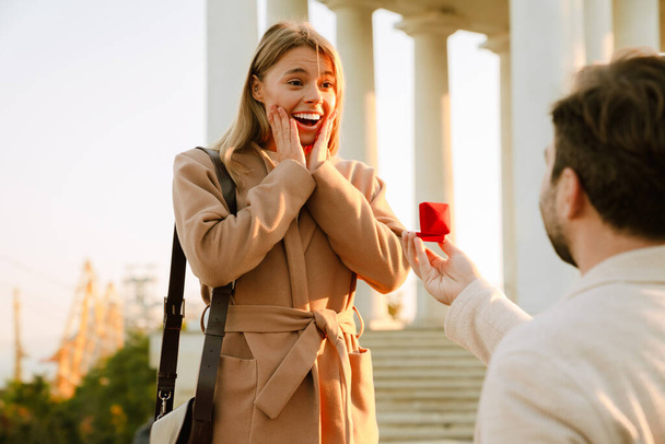 White man proposing to his excited girlfriend while standing on knee outdoors - Foto, Bild