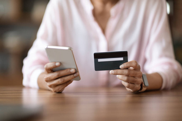 Przycięta kobieta w różowej bluzce siedząca przy biurku, trzymająca nowoczesną komórkę i plastikową kartę kredytową, nierozpoznawalna kobieta robiąca zakupy online lub płacąca rachunki przez Internet. Koncepcja bankowości internetowej - Zdjęcie, obraz