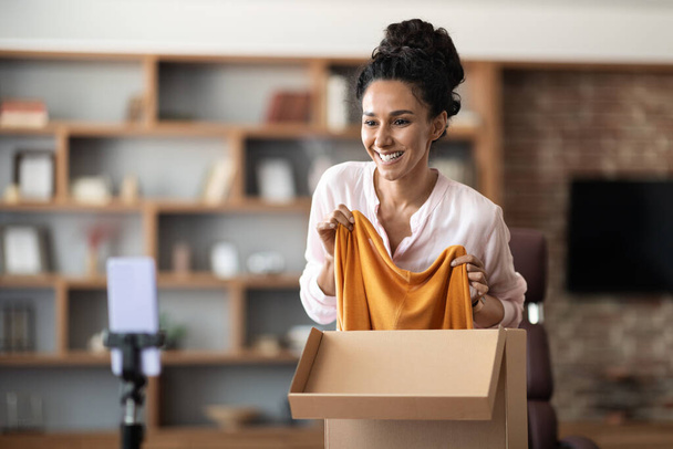Cheerful pretty young woman unpacking parcel and recording video, streaming via cell phone set on tripod, excited lady having video chat with friend, showing her new clothes and smiling, copy space - Φωτογραφία, εικόνα