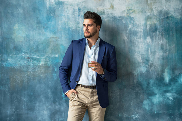 Young handsome businessman posing in elegant fashionable clothes, holding a glass of whiskey, tkinking and looking away. Portrait of bearded man. A lot of copy space on the background. - Foto, Imagem
