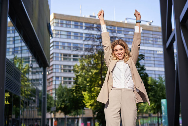 Portret van een gelukkige jonge zakenvrouw tilt de handen op, pompt vuist, viert overwinning, wint smth, bereikt doel, staat buiten op straat. - Foto, afbeelding