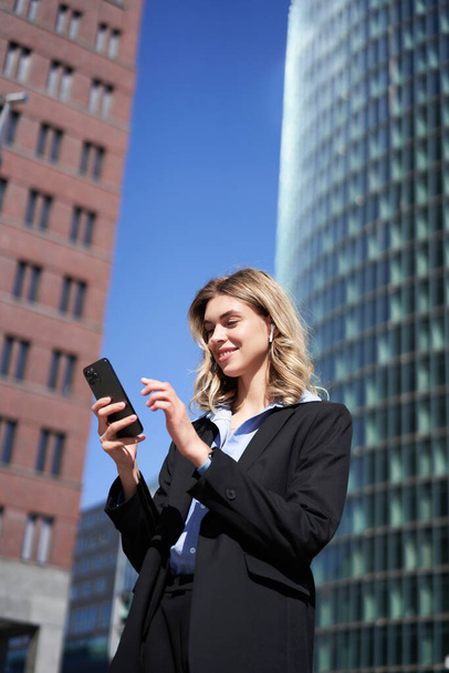 Portrait of successful businesswoman having a telephone conversation in wireless headphones, walking on street. Corporate woman with smartphone in city center. - Fotó, kép