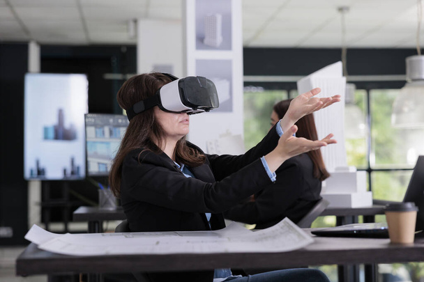 Architekt noszący zestaw słuchawkowy VR projektujący metawersy, dotykający wirtualnego modelu budynku, pracujący nad projektem. Inżynieria pracownicza w biurze architektonicznym, pracownik agencji nieruchomości w okularach ar - Zdjęcie, obraz