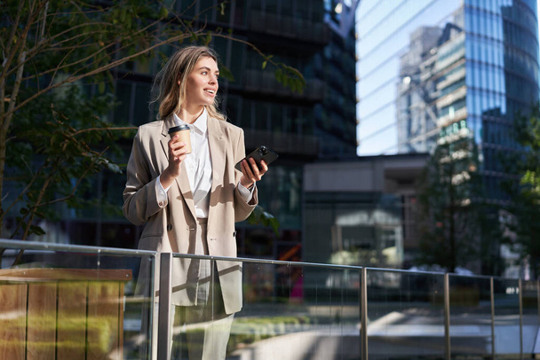 Successful businesswoman in suit, drinks her coffee and uses mobile phone, standing on street, having a lunch break. - Foto, Bild