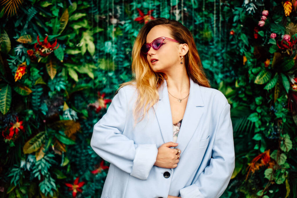 A girl in a dress, jacket and purple sunglasses poses at the festival portrait. Smile and joy, emotions against the backdrop of greenery and a waterfall. fashion outfit - Photo, Image