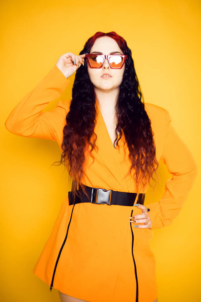 Bruneta v červených brýlích na oranžovém pozadí v kabátě. Módní fotografie modelu ve slunečních brýlích. - Fotografie, Obrázek
