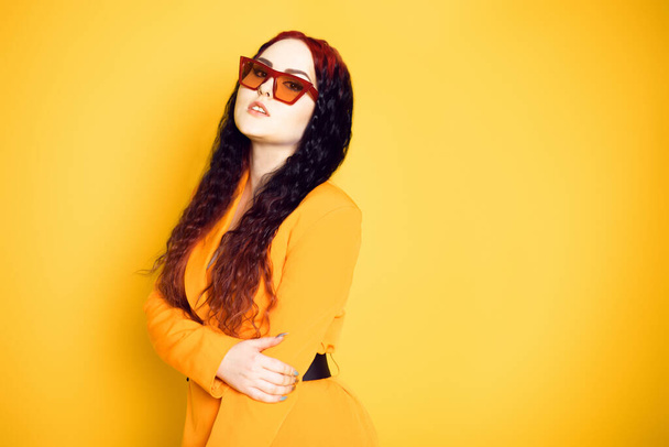 Brunette in red glasses on an orange background in a dress jacket. Fashion photo of model in sunglasses portrait - Photo, Image