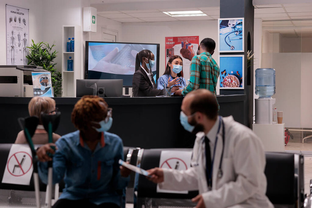 Női kórházi alkalmazottak, akik egy fiatal afro-amerikai férfit látnak el a recepción. Orvos átadja a receptet egy parciális test bénulásos nőnek. Orvosi klinika lobby alatt kovid - Fotó, kép