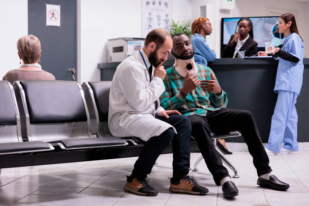 Médico de rehabilitación física que evalúa lesiones accidentales de varones afroamericanos jóvenes en la sala de espera del consultorio médico. Paciente explicando síntomas de lesión espinal. - Foto, Imagen
