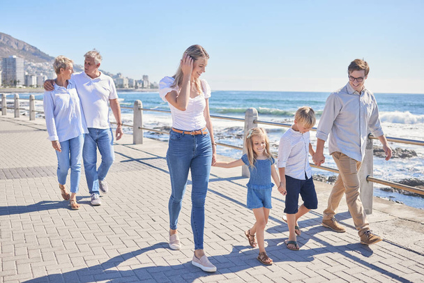 Loving caucasian parents holding hands with their little daughter and son while their grandparents follow in the background. Happy multi-generation family walking along seaside promenade on a sunny d. - Photo, Image