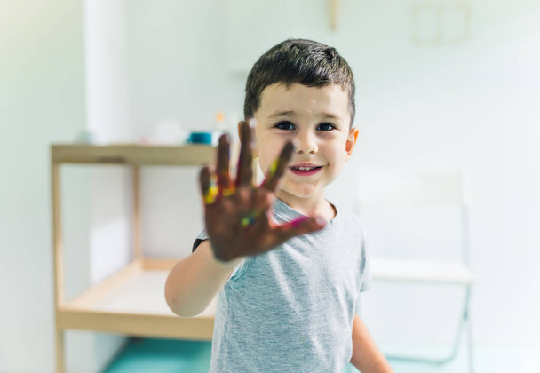 adorable niño pequeño con grandes ojos oscuros mostrando su mano pintada en la cámara, jardín de infantes de tiro medio. Foto de alta calidad - Foto, imagen