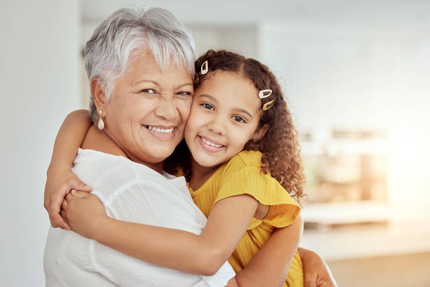 Portrait of mixed race grandmother and granddaughter hugging in living room at home. Smiling hispanic girl embracing senior grandparent and bonding in lounge. Happy elderly woman and child together. - Photo, Image