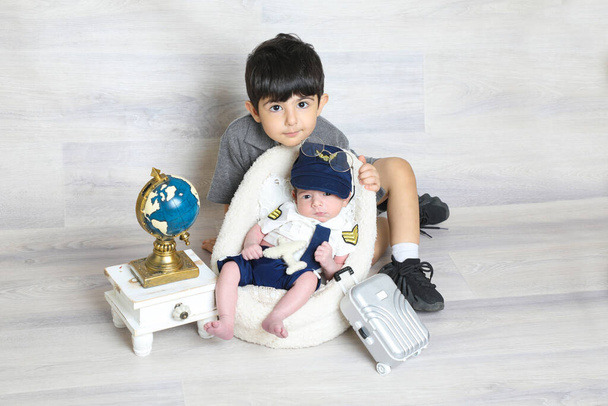 niño pequeño en uniforme piloto sentado en una silla pequeña y suave con su hermano mayor, maleta de juguete mapa del mundo fotografía de la cara niños árabes - Foto, imagen