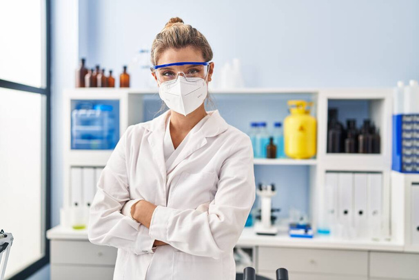 Молода блондинка в уніформі вченого і медична маска, що стоїть на руках, схрестила жест у лабораторії
 - Фото, зображення