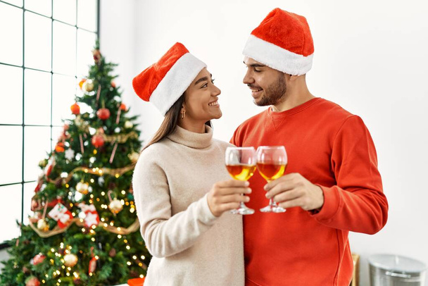 Young hispanic couple smiling happy toasting with champagne standing by christmas tree at home. - Photo, Image