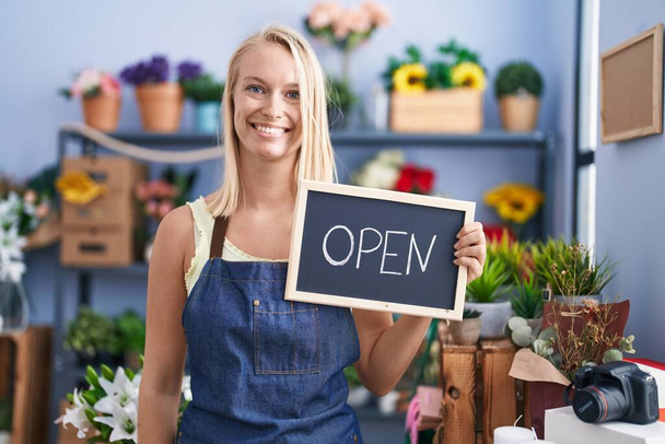 Young caucasian woman working at florist with open sign looking positive and happy standing and smiling with a confident smile showing teeth  - 写真・画像