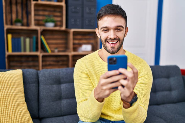 Fiatal spanyol férfi használ okostelefon ül kanapén otthon - Fotó, kép