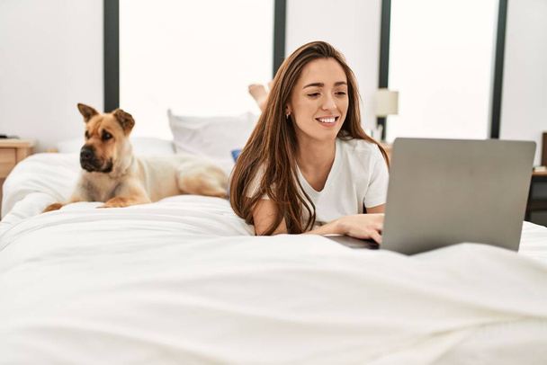 Young hispanic woman using laptop lying on bed with dog at bedroom - Foto, Imagen