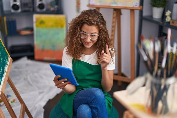 Genç beyaz kadın ressam, resim stüdyosunda oturmuş tabletle video görüşmesi yapıyor. Bir fikir veya soruyla gülümsüyor, mutlu bir yüzle işaret ediyor.  - Fotoğraf, Görsel