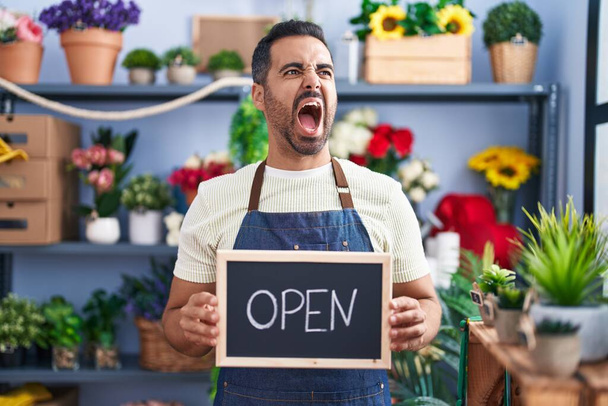 Hispanic man with beard working at florist holding open sign angry and mad screaming frustrated and furious, shouting with anger. rage and aggressive concept.  - Photo, Image
