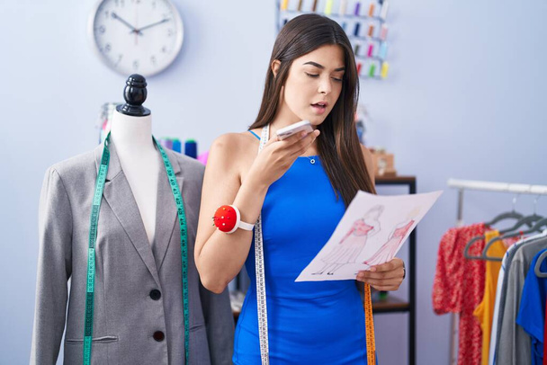 Jovem mulher hispânica bonita alfaiate falando no design de roupas procurando smartphone na loja de alfaiate - Foto, Imagem