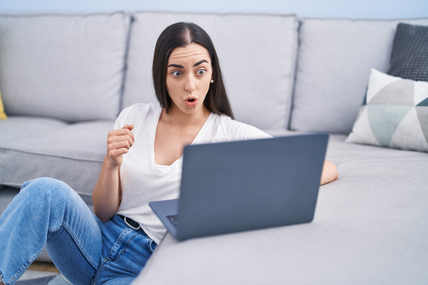 Nuori brunette nainen käyttää kannettavaa tietokonetta kotona peloissaan ja hämmästynyt suu auki yllätys, epäusko kasvot  - Valokuva, kuva