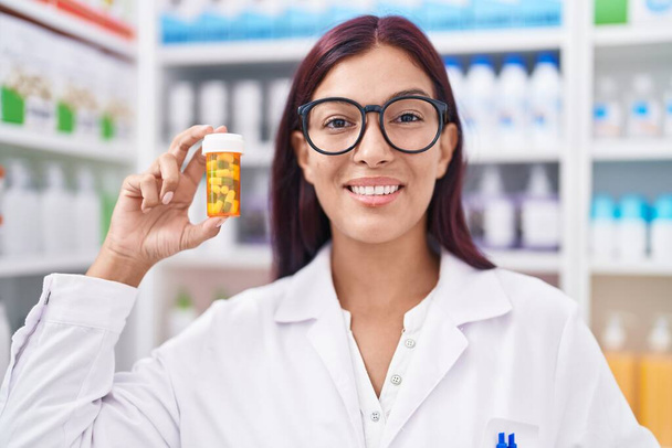 Giovane bella donna ispanica farmacista sorridente fiducioso tenendo pillole bottiglia in farmacia - Foto, immagini